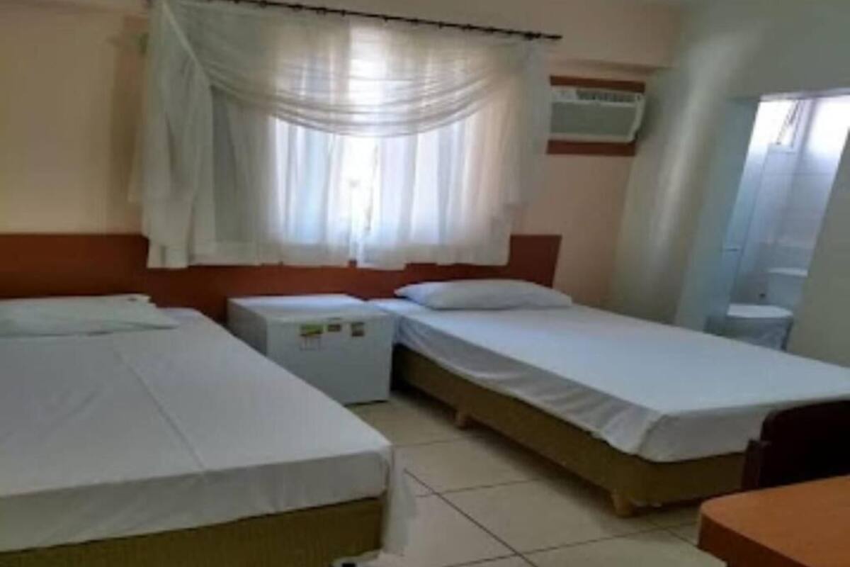 A room with two beds, a small table, a nightstand, and a window with sheer curtains.