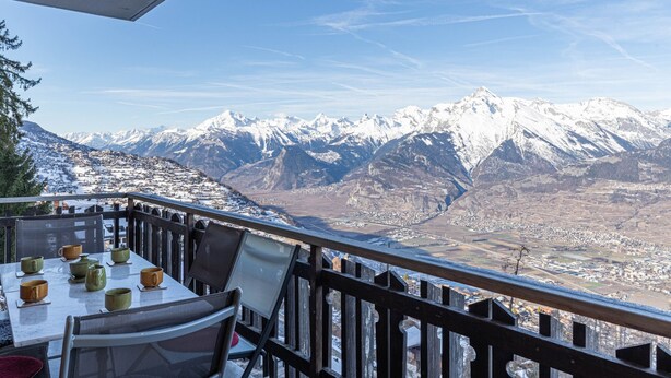 Ein Balkon mit Tisch für zwei Personen, mit Blick auf schneebedeckte Berge und ein Tal.