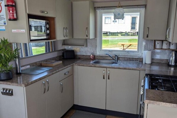 A kitchen with a microwave, sink, and stove. There is a window with a view of a house and a picnic table outside. - Cabin Sleeps 8 - Pools, Parking, Pets (Weymouth)