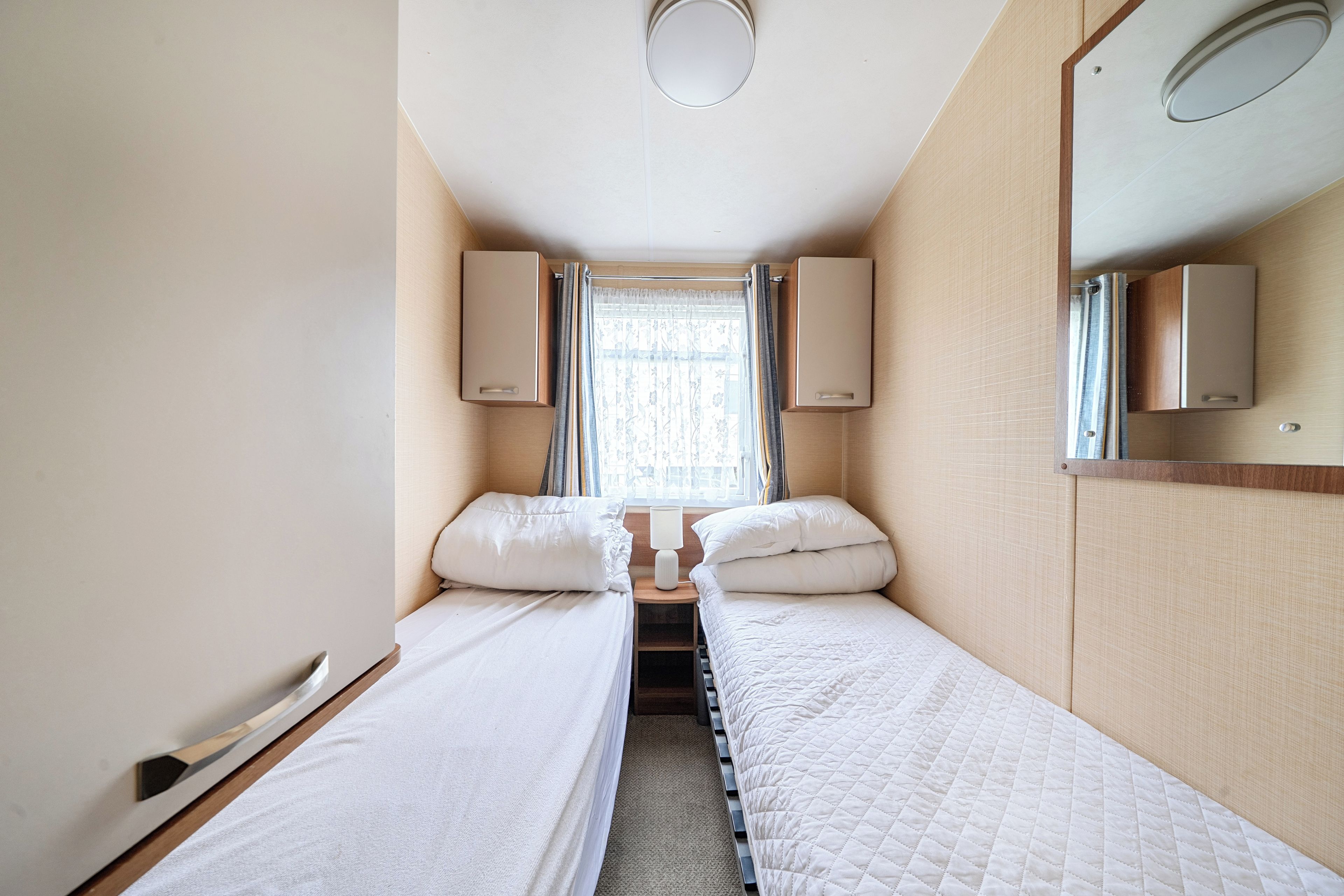 A narrow hotel room with two single beds, a window with curtains, and a mirror on the wall.