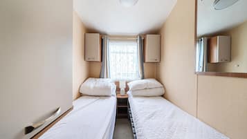 Une chambre d’hôtel étroite, avec deux lits simples, une fenêtre avec des rideaux et un miroir au mur.