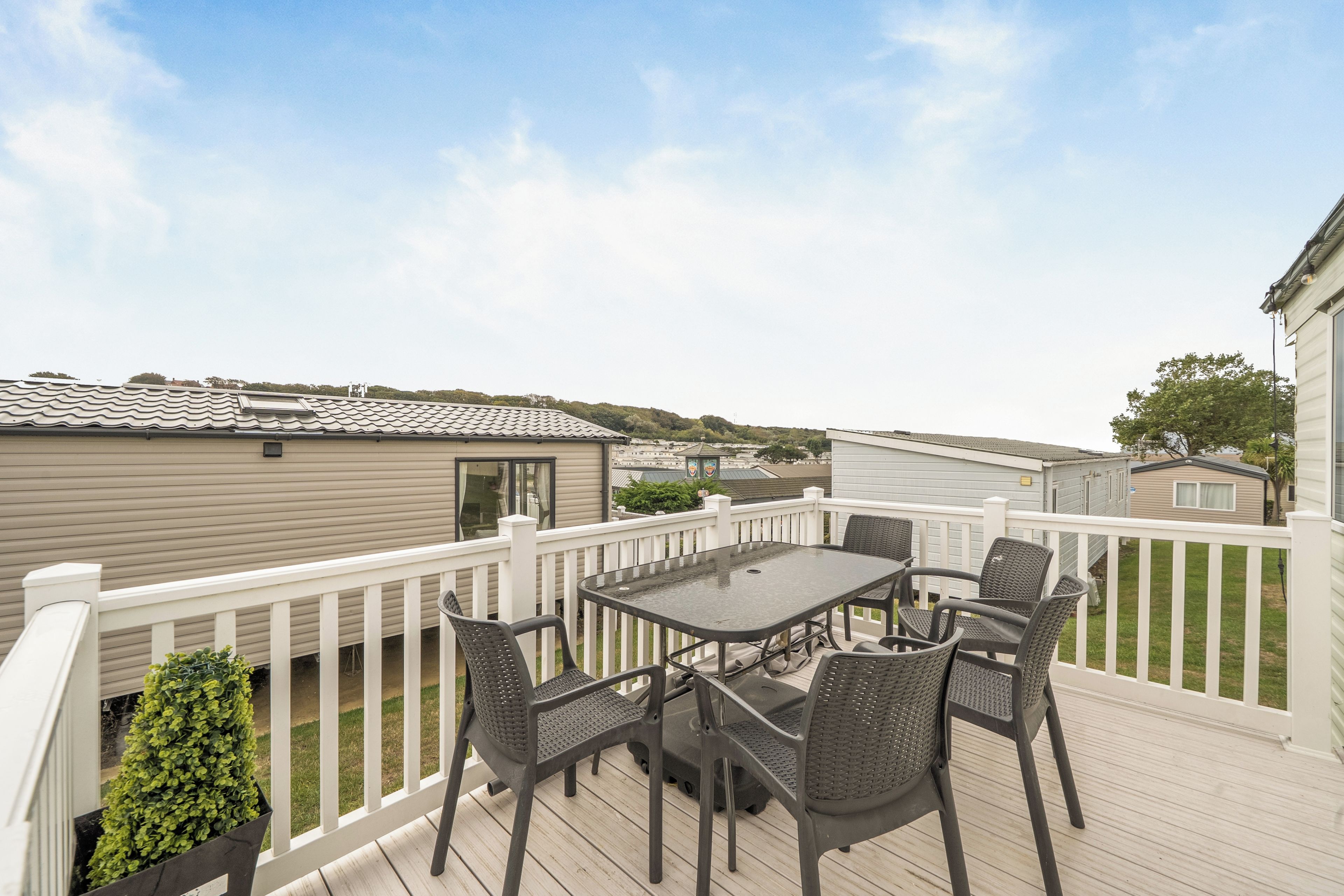 A deck with a table and chairs, surrounded by a white railing, overlooking a residential area with mobile homes.