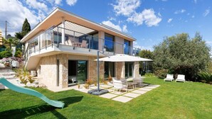 Una casa moderna de dos pisos con un balcón, un patio con muebles de exterior y un tobogán pequeño en el jardín.