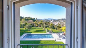 Blick von einem Balkon auf einen Pool, viel Grün und eine entfernte Stadtlandschaft.