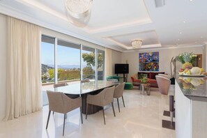 A modern living room with a dining area, large windows, a flat-screen TV, and a colorful painting.