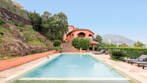 Piscina com borda de pedra, escadaria que leva a um edifício e cadeiras de descanso.