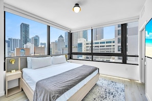 A modern hotel room with a large bed, a nightstand, a desk, and a view of the city skyline through large windows. - District 225 • 2 BD/2BTH • City Views • Free Spa (Miami)
