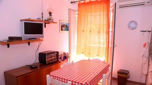 A room with a table covered by a checkered tablecloth, a television on a shelf, a clock on the wall, and a window with curtains. - Corallo (Tropea)
