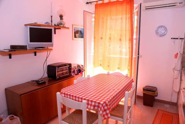A room with a table covered by a checkered tablecloth, a television on a shelf, a clock on the wall, and a window with curtains.