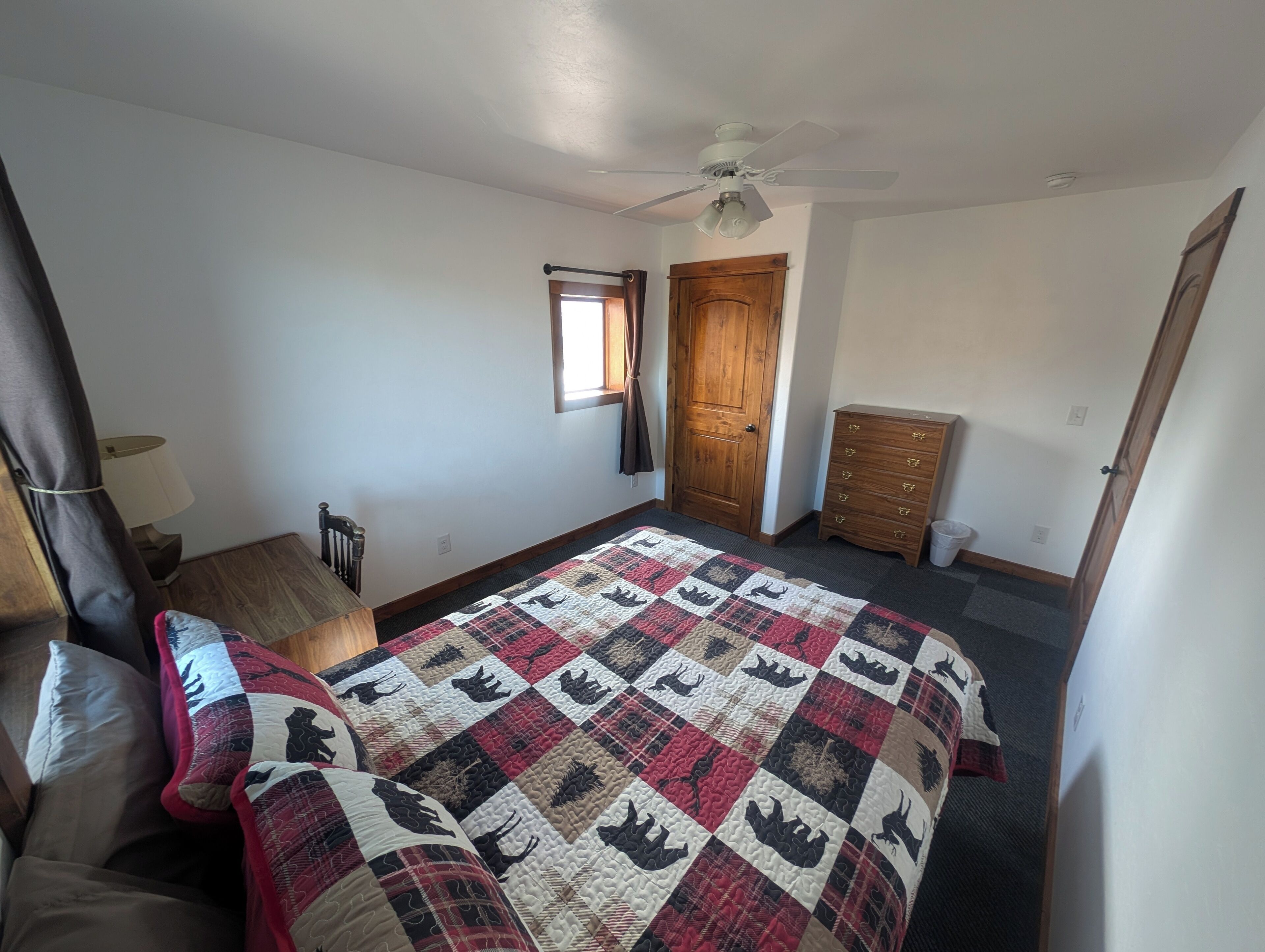 Een slaapkamer met een bed, een dressoir, een stoel, een raam met gordijnen, een plafondventilator en een deur.