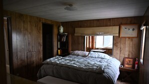 Quarto com paredes de madeira, cama com colcha, mesa de cabeceira com abajur e estante de livros.