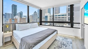 Ein modernes Hotelzimmer mit einem großen Bett, einem Nachttisch, einem Schreibtisch und Blick auf die Stadtlandschaft durch große Fenster.