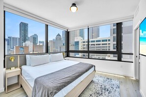 A modern hotel room with a large bed, a nightstand, a desk, and a view of the city skyline through large windows. - District 225 • 2 BD/2BTH • 20th Fl • Free Spa (Miami)