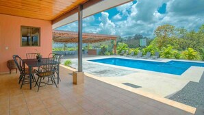 Ein Poolbereich im Freien mit einer überdachten Terrasse, einem Tisch mit Stühlen und Blick auf den Himmel und die umliegende Natur.
