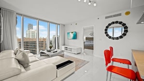 A modern living room with a white sofa, red chairs, a TV, and a large window with a city view. - Icon Brickell 2BR Ocean & Pool View + Spa Access (Miami)