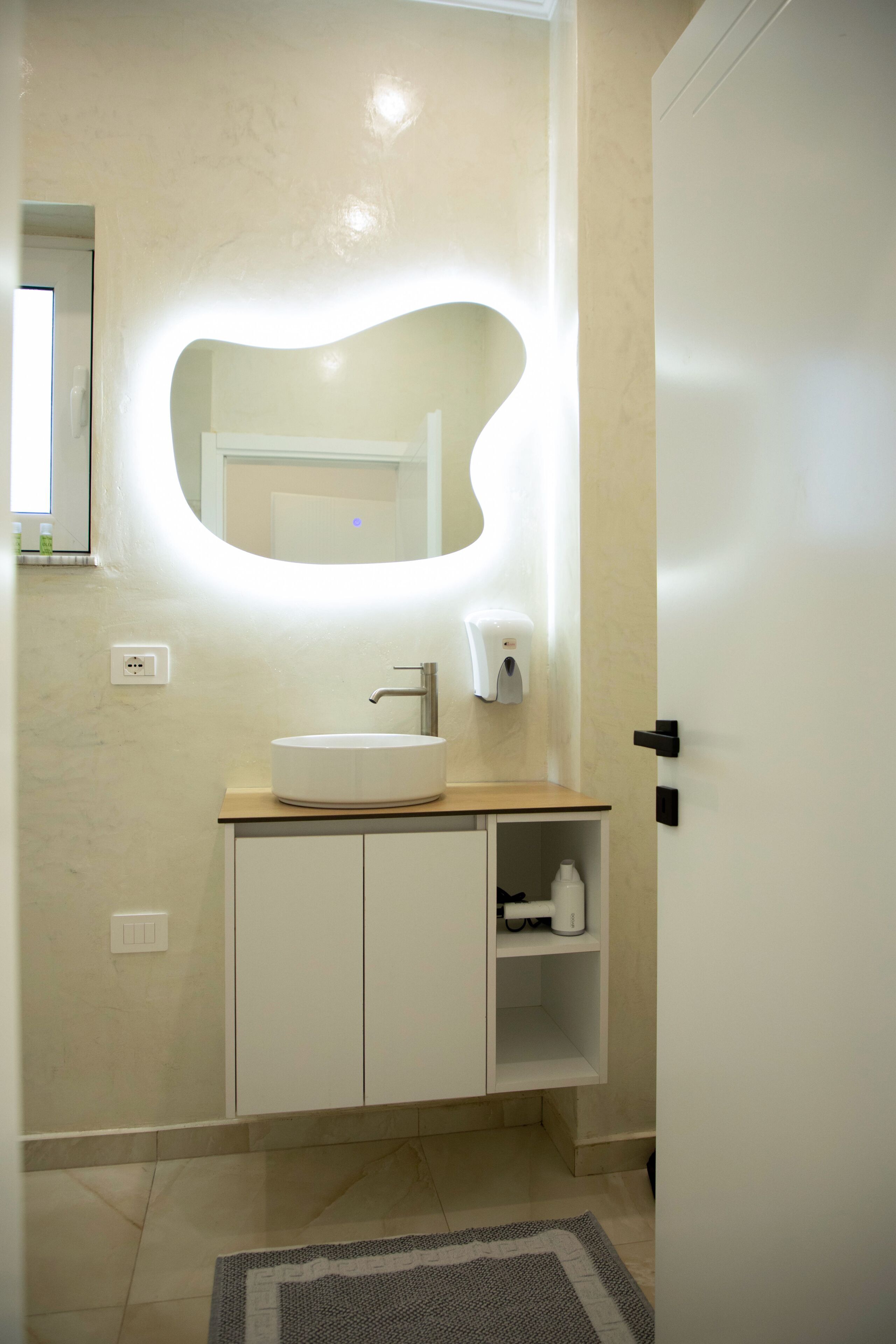 Un baño con un lavamanos redondo, un espejo, un dispensador de jabón y una puerta.