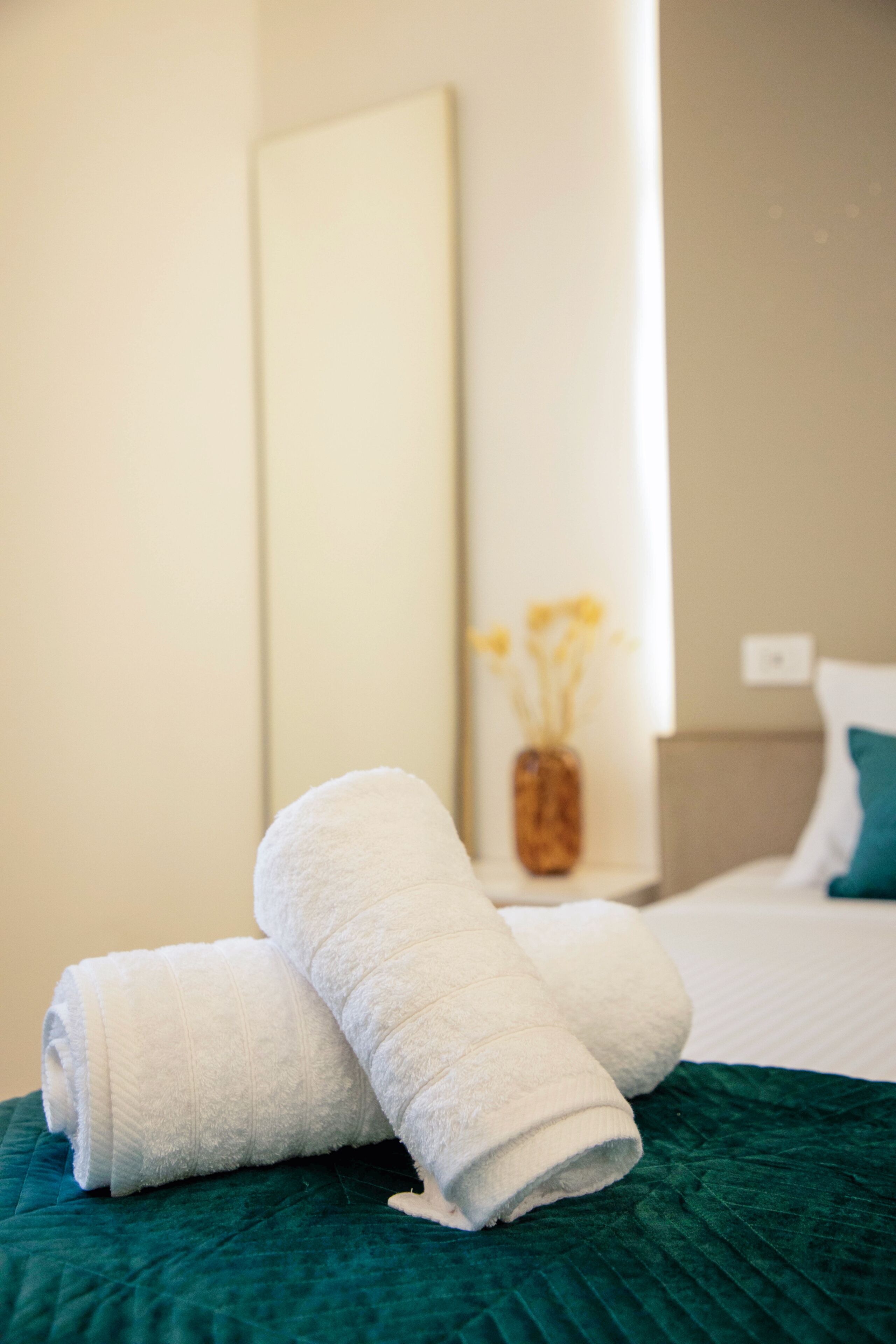 A neatly made bed with a green quilt, a white towel, and a vase with yellow flowers on a bedside table.