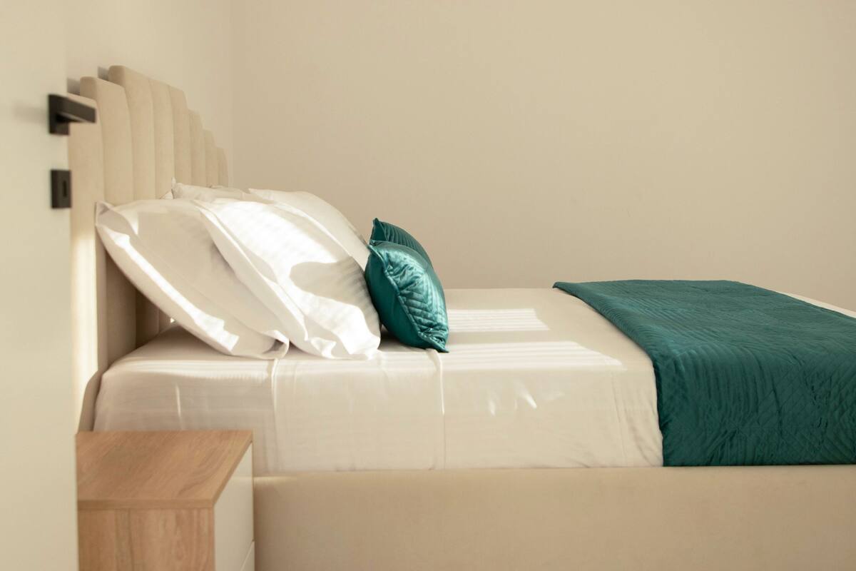 Un dormitorio moderno con cama, almohadas, una mesita de noche y una manta verde.