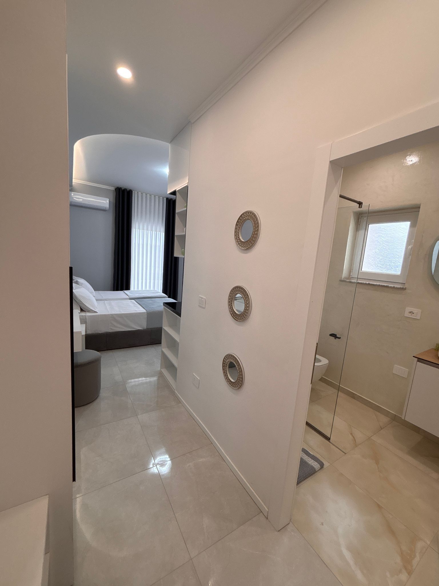 A modern hotel room with a bed, bedside tables, a mirror, and a small window.