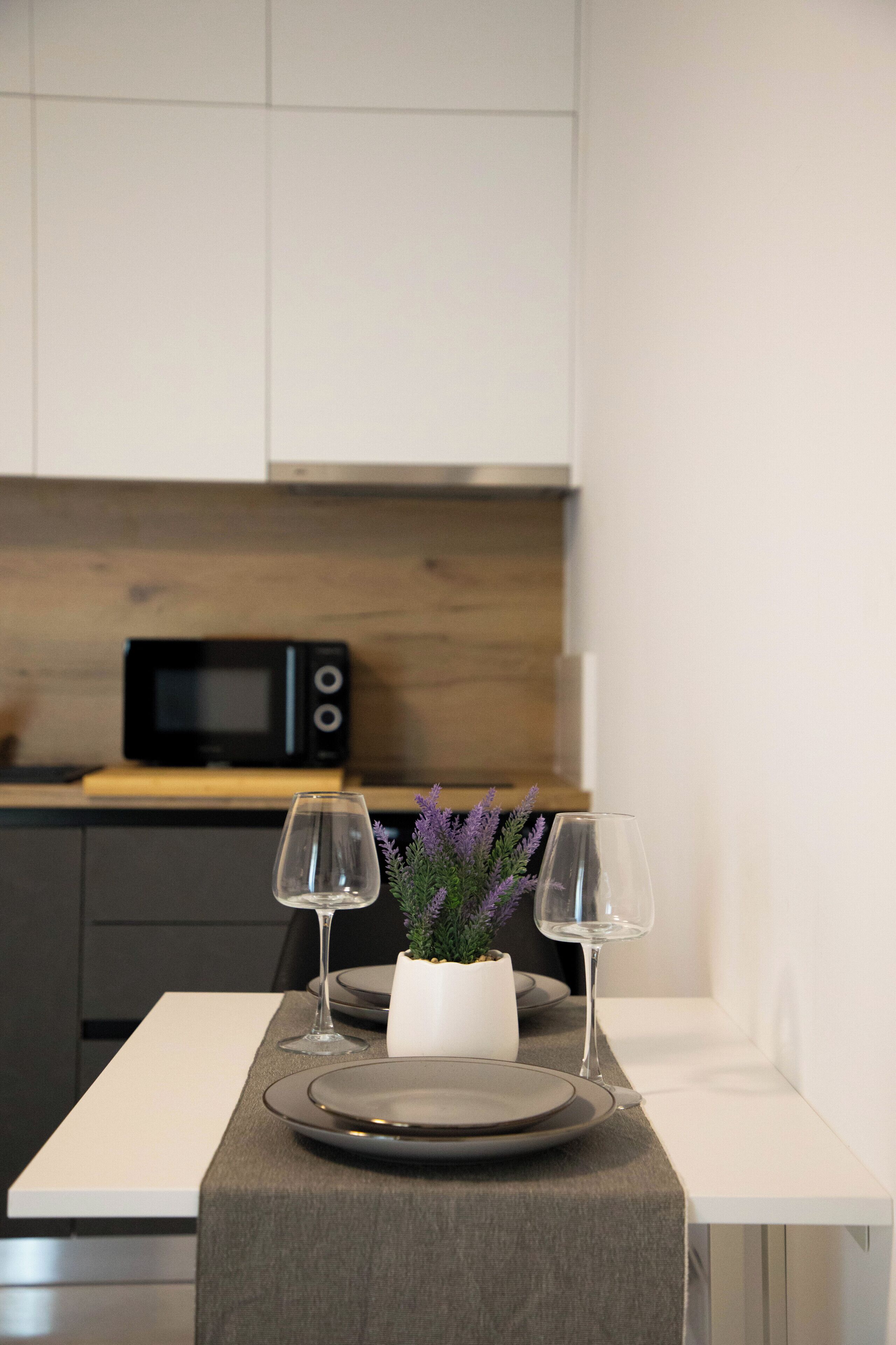 A modern kitchen with a microwave, a dining table set with plates and wine glasses, and a potted plant with purple flowers.