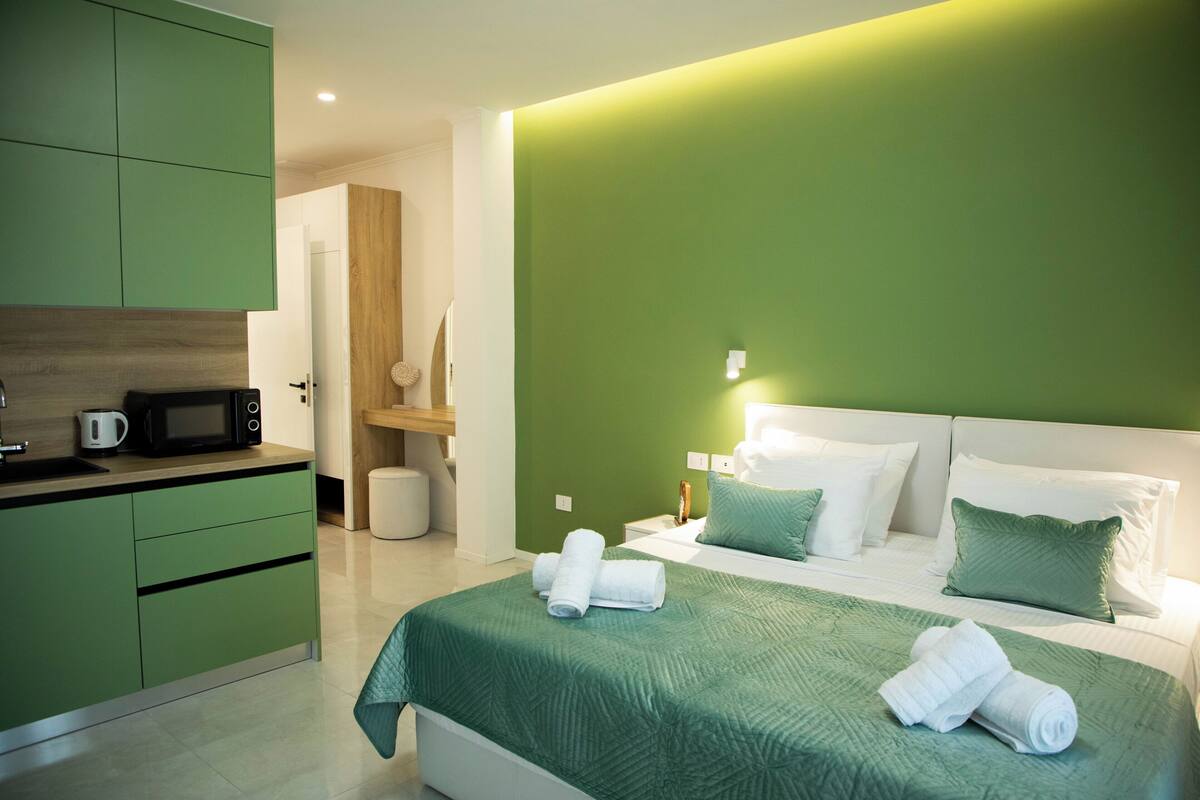 Una habitación de hotel moderna con una decoración verde, una cama con ropa de cama verde, un cabecero blanco y una zona de cocina con armarios verdes y un microondas.