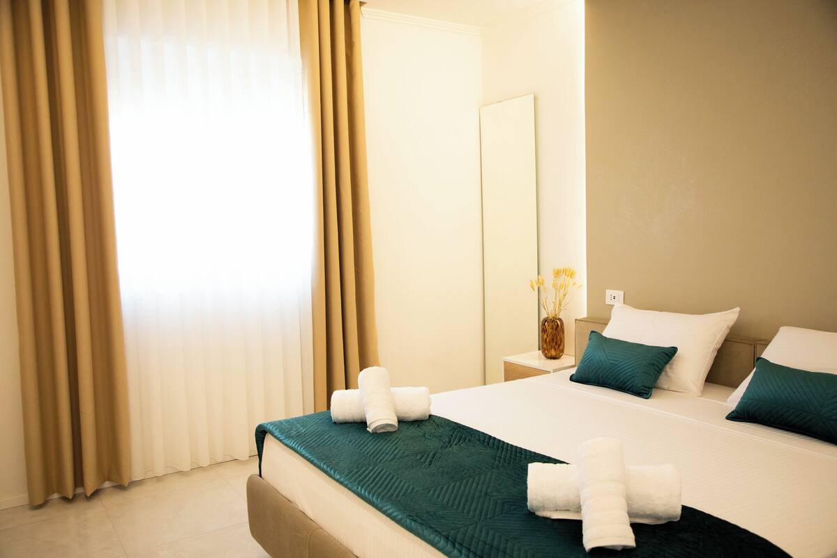 Una habitación de hotel con cama, mesita de noche, espejo y ventana con cortinas.