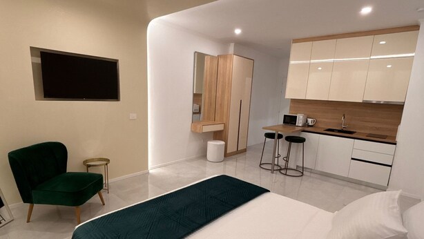 Une chambre moderne avec un grand lit, un fauteuil vert, un petit bureau et un coin cuisine.
