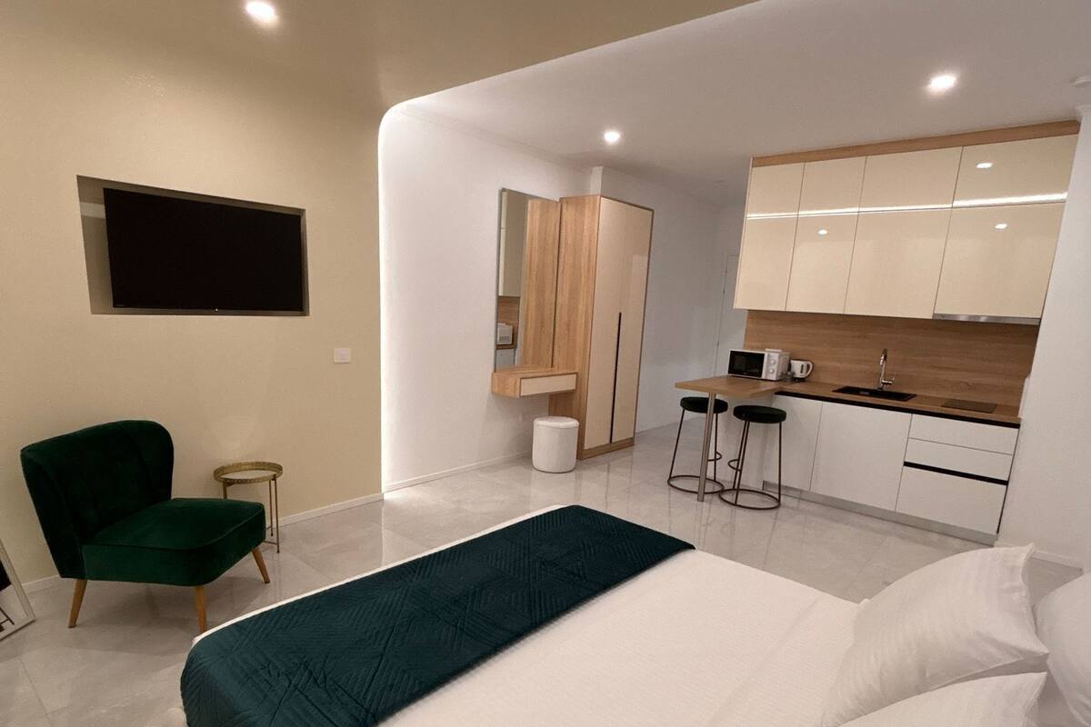 Un dormitorio moderno con una cama grande, una silla verde, un escritorio pequeño y una kitchenette.