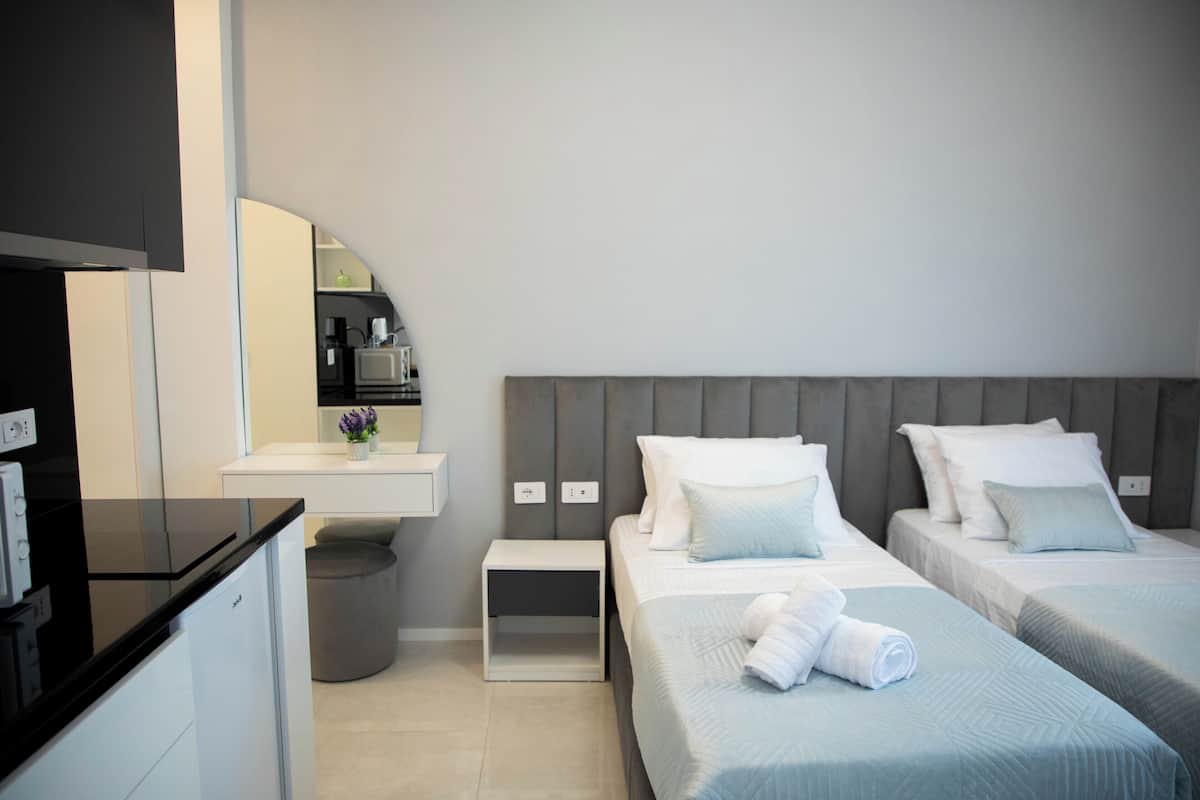 Una habitación de hotel moderna con kitchenette, baño pequeño y dos camas con ropa de cama blanca.