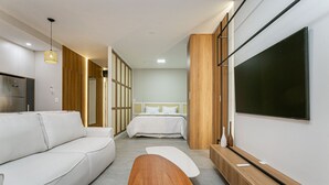 Et moderne hotelværelse med sofa, sofabord i træ, fjernsyn og seng med hvide sengetæpper.