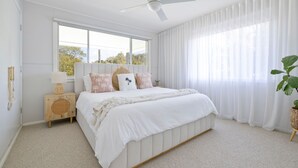Ein Schlafzimmer mit Bett, Deckenventilator, Fenster mit Vorhängen, Nachttisch mit Lampe und einer Pflanze.