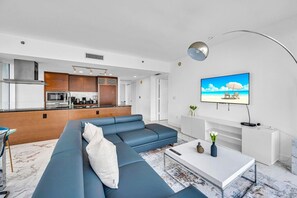 A modern living room with a blue sofa, a white coffee table, and a TV displaying a beach scene. - Icon Brickell 2BDR 44th Fl Ocean Views+Spa Access (Miami)