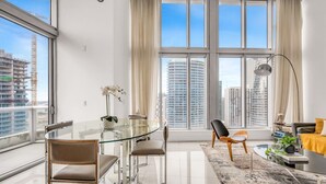 A modern living room with a glass dining table, two chairs, a sofa, a floor lamp, and a view of tall buildings. - Icon Brickell 2BDR 40th Fl Ocean Views+Spa Access (Miami)