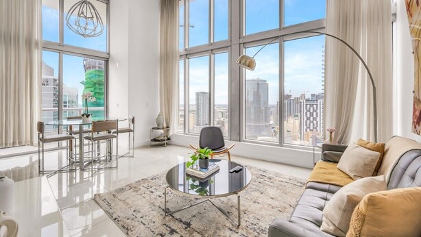 A modern living room with a glass coffee table, a sofa, a dining area with chairs, and large windows offering a city view. - Icon Brickell 2BDR 40th Fl Ocean Views+Spa Access (Miami)