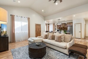 A living room with a sofa, ottoman, and round coffee table, a kitchen in the background, and a ceiling fan. - Welcome to Willow house. Your home away from home. (Pinetop-Lakeside)