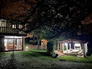 A nighttime view of a house with a covered patio, a visible staircase, and a well-lit interior.