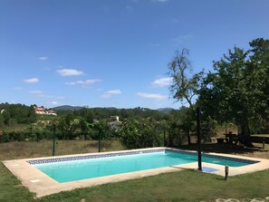 A rectangular swimming pool with a decorative border, surrounded by grass and trees, with a view of houses and hills in the background. - 'A Leira' House with Mountain Views, Private Pool and Garden (Ponteareas)