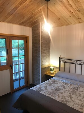 A bedroom with a wooden ceiling, a bed with a patterned bedspread, a nightstand with a lamp, and a view through a glass door. - 'A Leira' House with Mountain Views, Private Pool and Garden (Ponteareas)