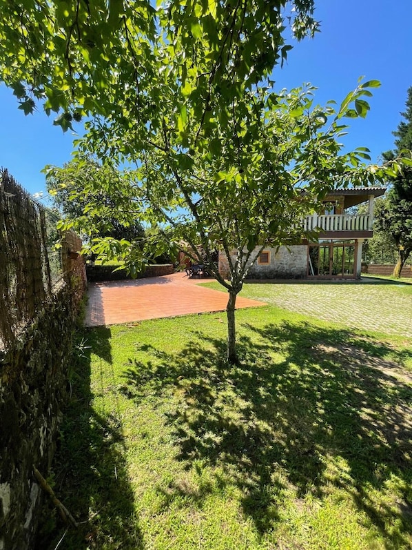 A tree-lined courtyard with a brick pathway, stone wall, and a building with a balcony.