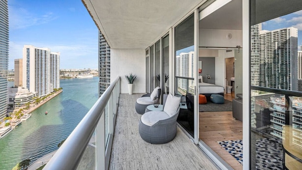 Ein Balkon mit Blick auf einen Fluss und hohe Gebäude, eine Sitzecke mit Kissen und ein modernes Interieur.