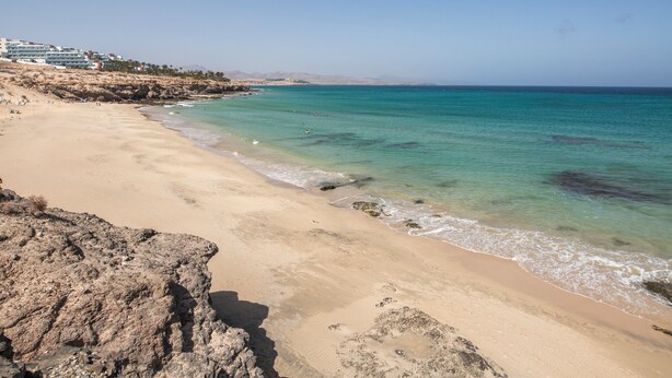 A beach with clear turquoise water, sandy shore, rocky outcrops, and a building complex in the background. - Heiwa Apartment with Shared Pool, Shared Garden, and Wi-Fi in Costa Calma (Costa Calma)