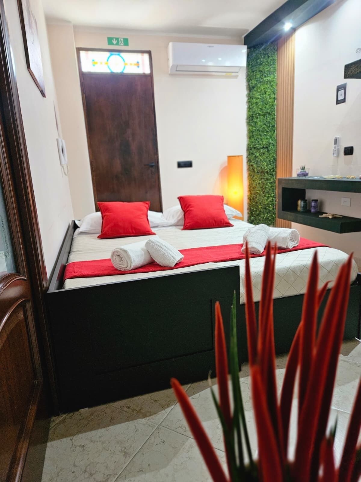 A hotel room with a bed, a wooden door, an air conditioning unit, a green wall, and a small shelf with toiletries.