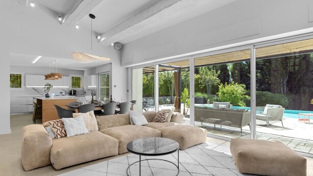 Ein modernes Wohnzimmer mit einem großen Ecksofa, einem runden Couchtisch und einem Essbereich, im Hintergrund ist eine Küche erkennbar.