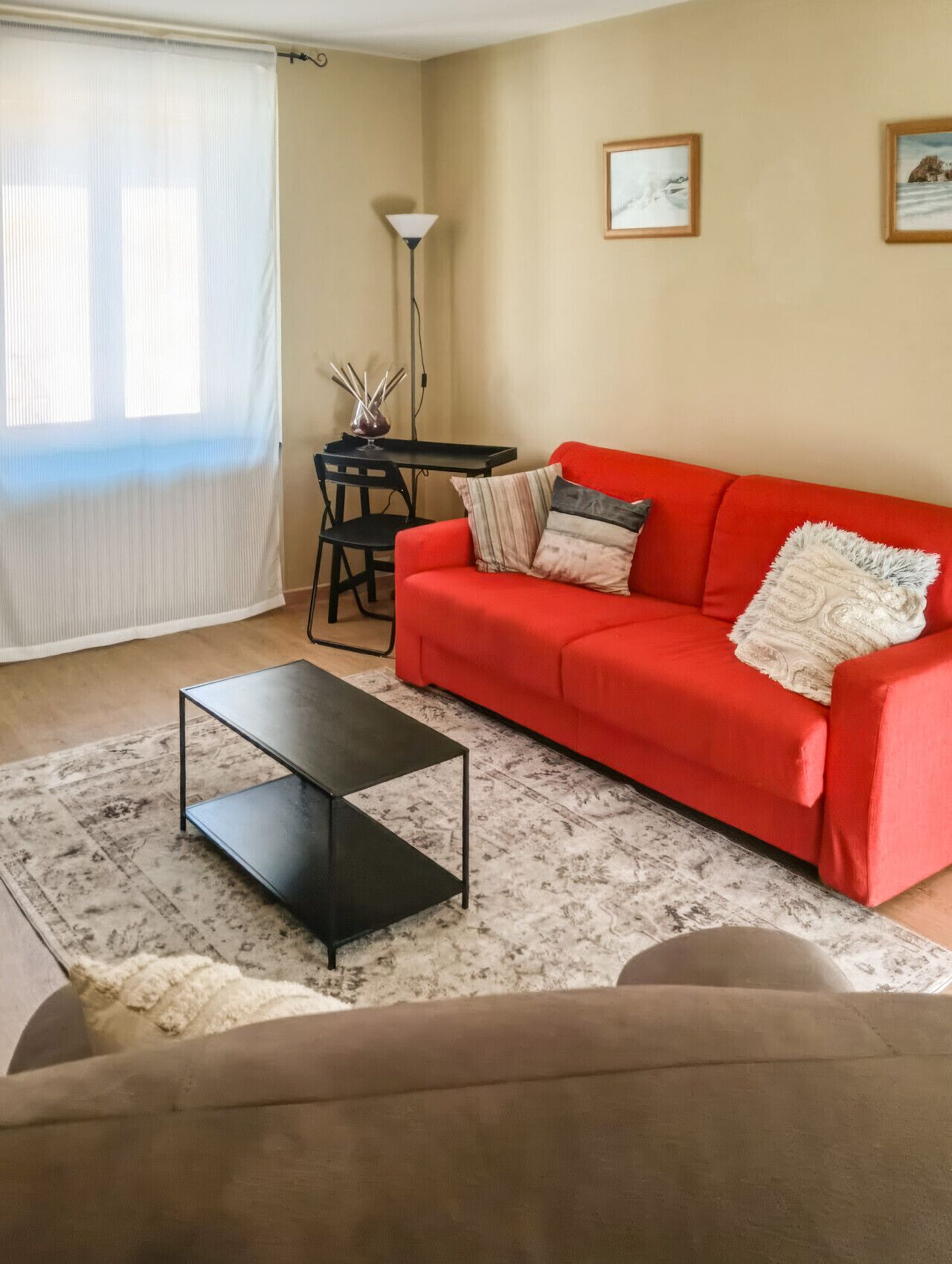 Un salon avec un canapé rouge, une table basse noire, une petite table avec une lampe et une fenêtre ornée de rideaux légers.