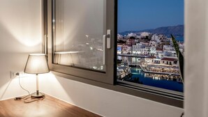 Une chambre avec vue sur une ville côtière la nuit, une lampe sur une table de chevet et une fenêtre donnant sur des bâtiments et l’eau.