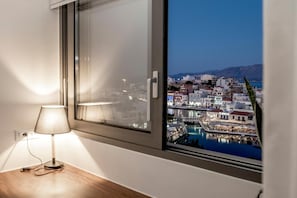A room with a view of a coastal city at night, a lamp on a bedside table, and a window with a view of buildings and water. - Apartment 'With Lake View, Jacuzzi' with Sea View, Wi-Fi and Air Conditioning (Agios Nikolaos)
