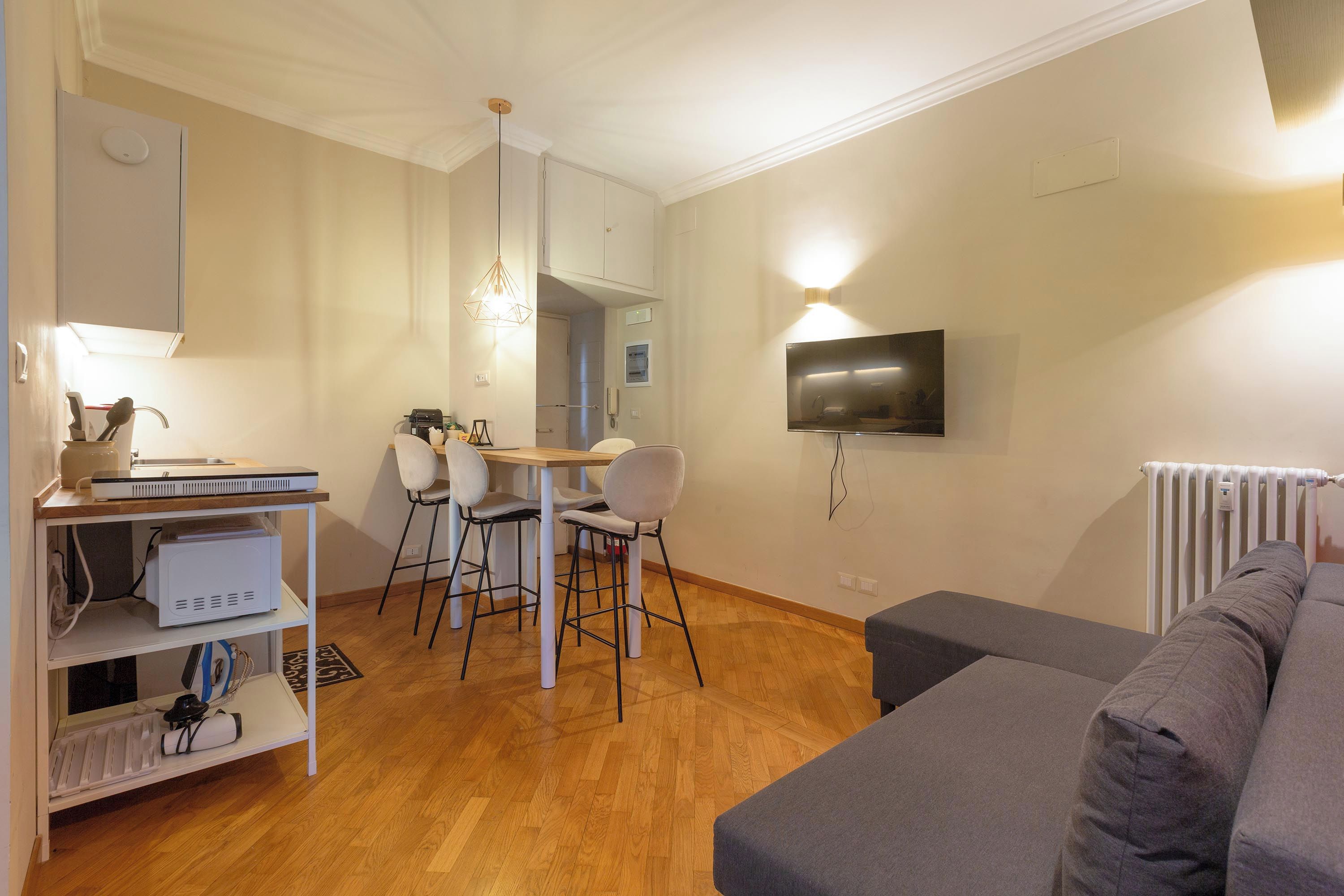 Un soggiorno moderno con un divano grigio, una zona pranzo con tavolo e sedie, un televisore fissato al muro e un termosifone.