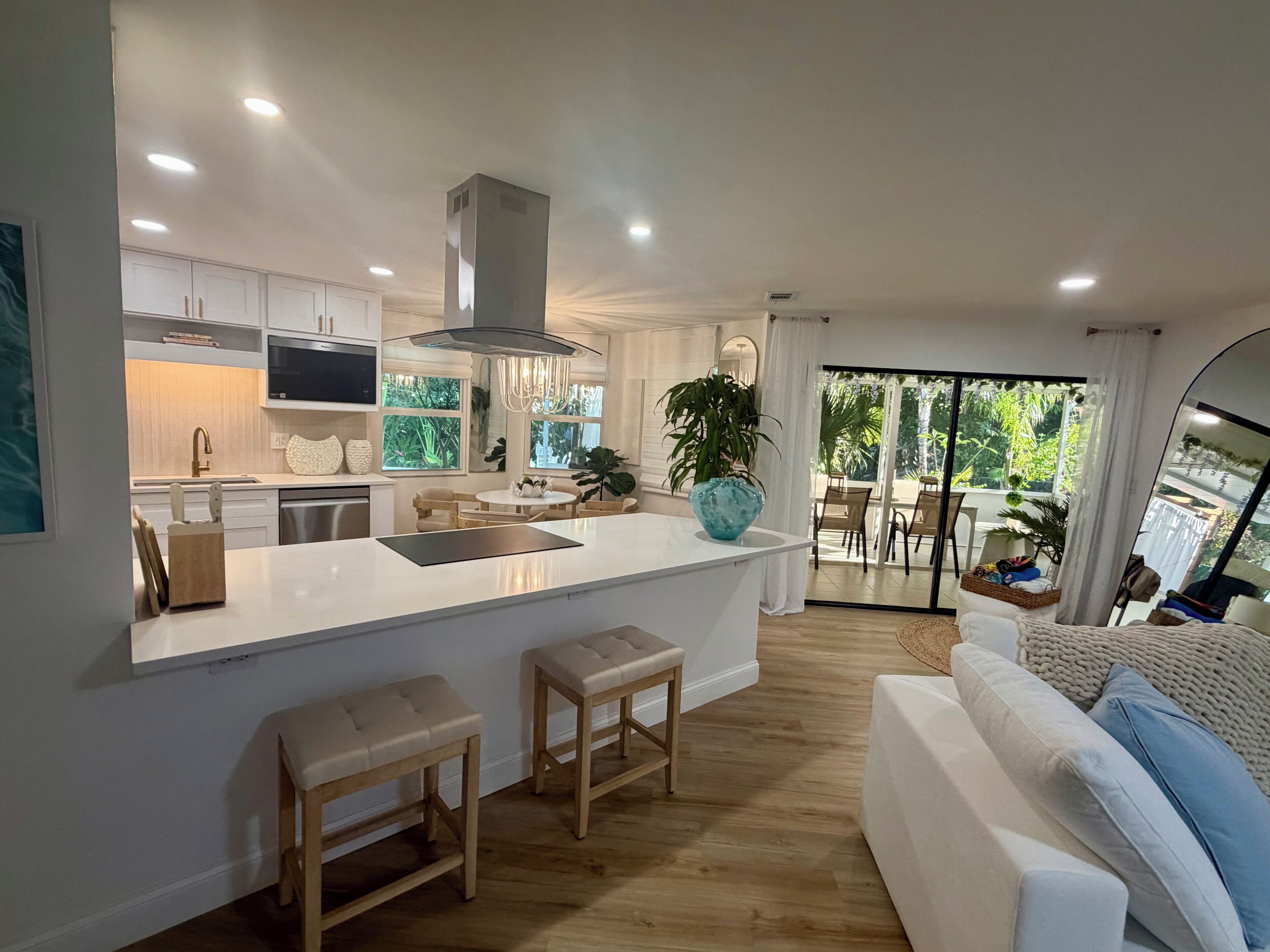 A modern kitchen with a central island, wooden stools, and a dining area with a view of the outdoors.
