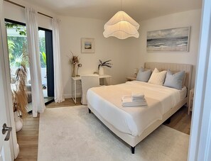 Un dormitorio con cama, mesitas de noche, un escritorio, una silla, una lámpara colgante, un cuadro y vistas a través de una ventana.