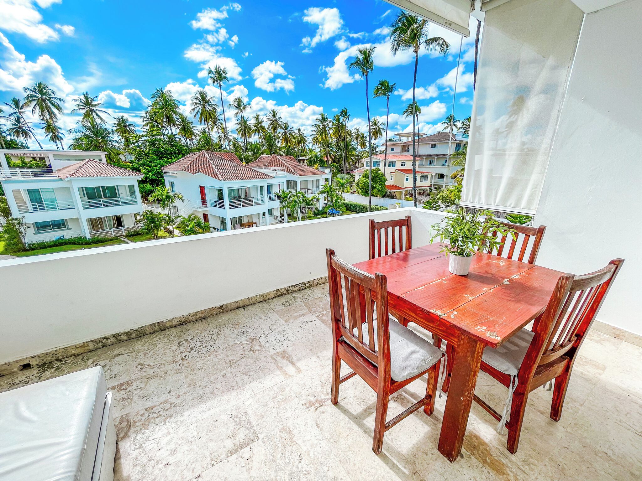 Un balcón con una mesa y sillas de madera, con vistas a una zona residencial con palmeras y casas.